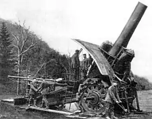 Photographie en noir et blanc représentant un groupe de cinq à six hommes servant une "Grosse Bertha". Les roues de l'obusier sont plus grandes qu'une hauteur d'homme, son canon se dresse à 80 degrés prêt à déverser ses obus de 420 millimètres.