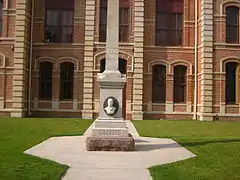 Visage sculpté à la base d'un poteau en pierre. En arrière-plan, la maison du comté.