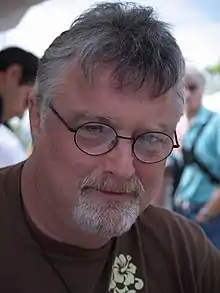 Homme avec des cheveux blancs et gris, des lunettes et un bouc.