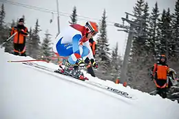 Skieur dans une descente, le corps groupé sur l'arrière des skis. Dans le fond, on a la présence de volontaires en veste rouge.