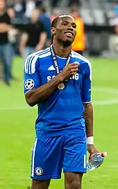 Photo en demi grandeur d'un footballeur en maillot bleu, une médaille au cou et une bouteille d'eau dans la main gauche