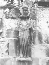 Photographie en noir et blanc d'une statue de pierre représentant un personnage debout et adossée à un mur.