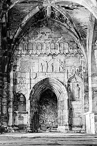 Photographie en noir et blanc du mur d'une église, percé d'une porte et sculpté d'une crucifixion et d'autres sujets.