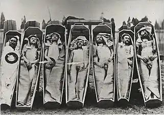 Photographie en noir et blanc représentant, en extérieur, sept cercueils sommaires dressés, contenant sept cadavres dont certains sont nus, d'autres recouverts d'un linceul ou drap. Les visages des cadavres restent visibles.