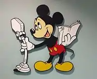 Le personnage Mickey chante avec un micro dans une main, et les paroles dans l'autre.