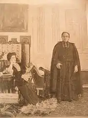 Photographie d'un prêtre en soutane et cape debout en conversation avec une dame assise dans un salon meublé bourgeoisement.