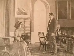 Une dame assise en conversation avec un homme debout devant elle dans une pièce meublée bourgeoisement avec un guéridon.