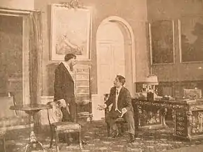 Un homme assis en conversation avec un autre homme barbu debout dans une pièce meublée bourgeoisement.