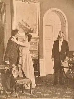 Une dame debout en conversation avec un homme debout devant elle et la femme suppliante pose sa main sur l'épaule de son interlocuteur dans une pièce meublée bourgeoisement. Un deuxième homme barbu les regarde surpris.