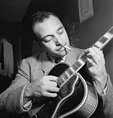 Django Reinhardt avec une guitare électrique entre les doigts, une cigarette à la bouche. Ses yeux sont tournés sur le manche de sa guitare. La photo est en noir et blanc.