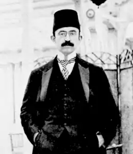 Portrait d'un homme moustachu portant un fez, un costume, une cravate et des lunettes.