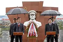 Photo en couleur de quatre hommes, deux derrière des podium, les deux autres tenant des parapluies, se tenant devant un monument portant un buste de Joukov.