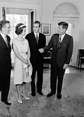 Photographie en noir et blanc montrant une conversation entre trois hommes portant un costume et une femme portant une robe.