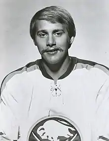 Portrait photographique noir et blanc d’un homme, de buste, moustachu, portant un maillot de hockey sur glace clair