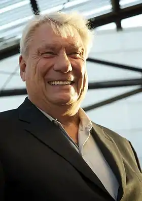 A man, wearing a brown coat, white shirt and black tie, standing in front of a collage.