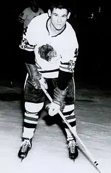 Photographie en noir et blanc d'un joueur de hockey sur glace posant avec un maillot frappé d'un tête d'un amérindien.