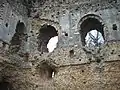 Photographie du coin d'un château fort depuis l'intérieur. Trois fenêtres sont visibles. Les murs sont en mauvais état, une grande partie du parement a disparu.