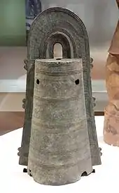 Photo couleur d'une cloche en bronze de forme cylindrique posée sur un support blanc.