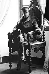 Un homme est assis dans une chaise richement décorée. Il porte un képi, un manteau et des bottes de cavalier et il tient une cravache.