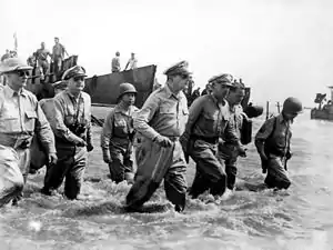 Un groupe d'hommes en uniforme progresse dans un mètre d'eau. Des péniches de débarquement sont visibles à l'arrière-plan