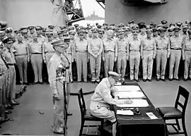 MacArthur est assis et écrit sur un petit bureau. Deux hommes en uniforme se tiennent derrière lui et une large foule d'hommes en uniformes observe la scène.