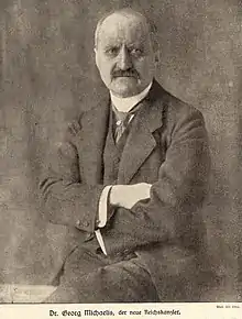 Photo en noir et blanc d'un homme âgé assis en costume les mains croisées, il porte une moustache et a le crane dégarni