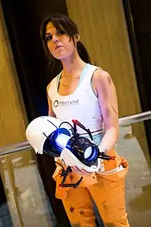 Femme d'environ 30 ans habillée avec un pantalon orange et un marcel blanc sérigraphié Aperture en noir tenant le Portal Gun dans ses mains.