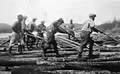 Photographie en noir et blanc avec des hommes debout sur des billots de bois flottant sur l'eau. Ils dirigent les billots avec de longues perches.