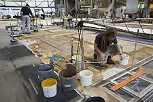 Dans un immense atelier, quelqu'un dessine avec soin une colonne au milieu des pots de peinture.