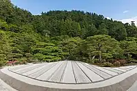 Jardin zen de sable sec à longues rayures beiges, vu de face avec une impression de perspective centrale, des fleurs et des arbres en arrière-plan, un jour ensoleillé au temple bouddhiste zen Higashiyama Jisho-ji, du Ginkaku-ji, le temple du Pavillon d'argent (juin 2019).