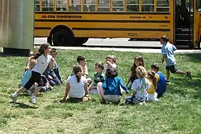Un groupe d'enfants sont assis en cercle dans l'herbe, et deux autres courent l'un après l'autre autour du cercle.