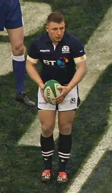 Photographie d'un joueur de rugby vêtu d'un maillot bleu foncé, d'un short blanc et porteur d'un ballon de rugby.