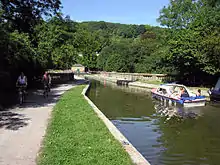 Le canal passant sur un pont avec des murs en pierre, entouré d'arbres. Sur la gauche et la droite se trouvent des chemins de halage avec les cyclistes et les piétons.