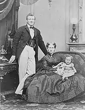 Photographie en noir et blanc d'un couple avec un bébé. Le père est debout et porte une veste foncée et un pantalon blanc, la mère est assise sur un petit divan et tient un bébé sur les genoux.