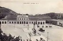 Carte postale en noir et blanc d'un bâtiment horizontal avec un grand parvis devant sur lequel se trouvent des calèches.