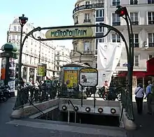 Photographie en couleurs d'une bouche de métro, avec portique en fer frogé très large et perspective diagonale d'une rue se prolongeant vers le fond de l'image