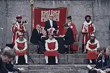 Photographie d'une estrade (sur des marches) où un homme en jacquette s'adresse à une assemblée ; autour de lui plusieurs personnes en différents costumes rouges et blancs ; au mur un drapeau affichant une double-clef blanche sur fond rouge.