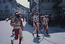 Photographie de trois hommes, portant la barbe et un costume en rouge et blanc, marchant dans une rue.