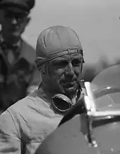 Photo en plan américain de Francis Curzon, en tenue de pilote et au volant d'une voiture.