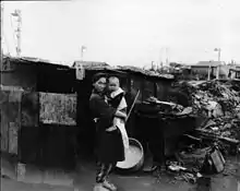 Photographie en noir et blanc d'une femme avec un habit traditionnel japonais portant un petit enfant devant une baraque hâtivement construite. Des tas de décombres et des maisons intactes sont visibles à l'arrière-plan.