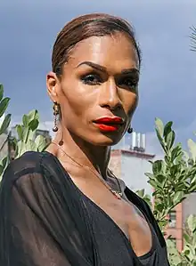 Photographie d'une femme Noire portant du maquillage sophistiqué, en particulier un rouge à lèvres intense, des bijoux dorés et un haut noir semi-transparent