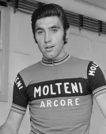 Photographie en noir et blanc d'un cycliste, debout devant un mur blanc et portant un maillot avec l'inscription Molteni Arcore.