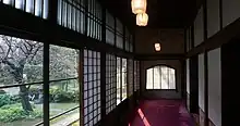 Un couloir de bois en enfilade, portes coulissantes en bois et papier sur la droite, fenêtres sur la gauche. tapis rouge au sol. À l’extérieur par les fenêtres, vue sur un  jardin avec un cerisier.