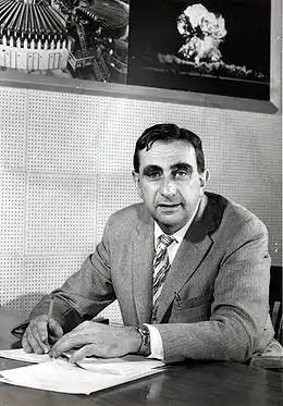 Photo en noir et blanc. Un homme portant un complet est assis à une table de travail. Derrière lui, deux photos sont accrochées au mur, dont l'une montrant un champignon atomique.