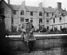Édouard VII portant un kilt et une canne est assis sur un muret devant une maison à trois étages.