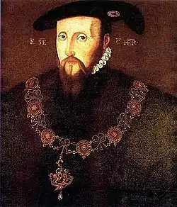 Portrait d'un homme à la barbe rousse portant un manteau et un chapeau noir ainsi qu'un collier de pierres précieuses