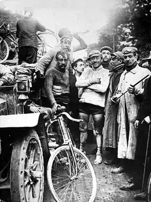 Photographie en noir et blanc d'un groupe d'hommes, parmi lesquels des cyclistes et des spectateurs, regroupés autour d'une voiture.