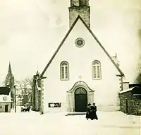 Une façade d'une petite église en plein hiver avec deux personne marchant devant en manteaux.