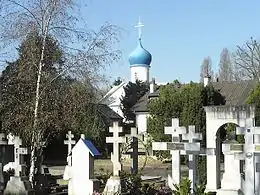 Photographie en couleur de tombes surmontées de croix russes avec, en dernier plan, une église russe avec un bulbe bleu.