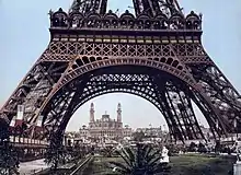 Photographie colorisée de 1900 pendant l'exposition, présentant une vue du palais sous les arches de la tour Eiffel.